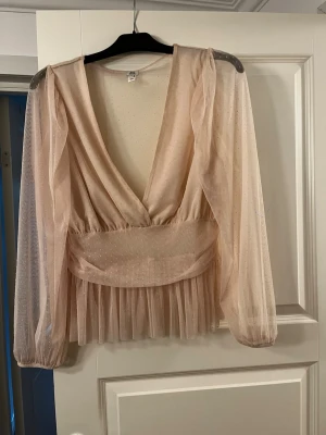 Beige meshblus med djup v-ringning - Snygg beige blus i transparent mesh med diskreta prickar, djup v-ringning och markerad midja. Långa puffiga ärmar och volang nedtill ger en trendig och romantisk look. Perfekt att styla med jeans eller kjol.