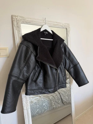 Svart skinnjacka med fuskpälskrage - Snygg svart skinnjacka från H&M med bred fuskpälskrage och dragkedja framtill. Jackan har två snedställda fickor och en oversized passform som ger en cool look. Perfekt för dig som vill ha en trendig och varm jacka till hösten.