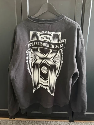 Svart sweatshirt från DOLLY NOIRE - Cool svart sweatshirt från DOLLY NOIRE med stort grafiskt tryck och texten 'Established in 2013' på ryggen. Liten logga på bröstet. Klassisk rund hals och ribbade muddar vid ärmar och nederkant. Perfekt för en avslappnad streetwear-look. Ny kostar mellan 1000-2000kr. Den sitter som L och den är lite sliten på trycket.