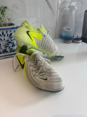 Nike Phantom GT2 fotbollsskor gul/grå - Säljer ett par Nike Phantom GT2 fotbollsskor i grått och neongult med svarta detaljer. Skorna har snörning, låg profil och mönstrad ovandel för bättre bollkontroll. Perfekta för dig som vill sticka ut på planen och få bra grepp på gräs. Du får även med kolfiber sulor som gör att du får bättre acceleration och kan hoppa högre
