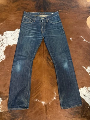 Nudie Jeans RegularRalf RegularAlf vintage 2000s bootcut - Säljer ett par klassiska mörkblå jeans från Nudie Jeans med raka ben och snygga slitningar. Jeansen har fem fickor, orange sömmar och Nudie-logga på bakfickan. Tillverkade i kraftig denim som ger en tidlös look.