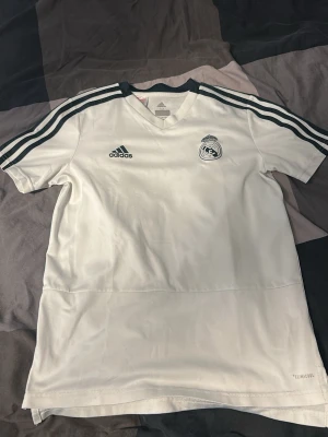 Vit Real Madrid tröja från Adidas - Snygg vit Real Madrid tröja från Adidas med svarta detaljer och klassiska ränder på axlarna. Tröjan har klubbmärket på bröstet och Adidas-logga. Tillverkad i ett lätt och funktionellt material, perfekt för träning eller match. Det står att det är M men den är 146.