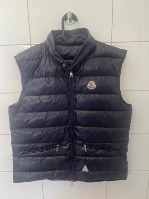 Mörkblå dunväst från Moncler - Snygg mörkblå dunväst från Moncler med klassisk quiltad design och hög krage. Västen har två dragkedjeförsedda fickor framtill och Monclers ikoniska logotyp på bröstet. Tillverkad i glansigt material med dunfyllning. Nfc scan✅ storlek 3 sitter som L