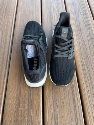 Adidas Ultra Boost svarta sneakers - Snygga Adidas Ultra Boost sneakers i svart med stickad ovandel och vita Boost-sulor. Skorna har klassiska tre ränder på sidan, gråa skosnören och en bekväm, sportig design. Perfekta för dig som gillar streetwear och sportig stil.