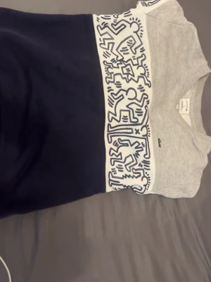 Lacoste x Keith Haring t-shirt grå/blå - Grå och marinblå sweatshirt från Lacoste x Keith Haring med ikoniskt grafiskt tryck i vitt och mörkblått över bröstet. Tröjan har rund halsringning och klassisk passform. Perfekt för dig som gillar streetwear och konstnärliga detaljer. Kan sänka pris vid snabb affär. Minimal skada vid arm änden 