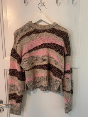 Stickad tröja med breda ränder - Mysig stickad tröja med breda ränder i beige, rosa och mörkbrunt. Tröjan har avslappnad passform, ribbade muddar och rund hals. Perfekt för dig som gillar en unik och färgglad look. Materialet känns mjukt och varmt.