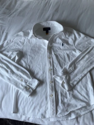 Vit skjorta från Polo Ralph Lauren - Klassisk vit skjorta från Polo Ralph Lauren med broderad logga på bröstet. Skjortan har lång ärm, knappar framtill och är tillverkad i mjuk bomull. Perfekt till en clean och stilren look.