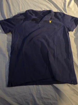 Blå pikétröja från Lyle & Scott - Mörkblå pikétröja från Lyle & Scott med klassisk krage och korta ärmar. Tröjan har en gul broderad logga på bröstet och är tillverkad i mjuk bomull. Perfekt för en avslappnad och stilren look.