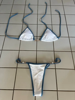 Vit bikini med blå kanter och pärldetaljer - Trendig bikini i vitt med blå kanter och snygga pärldetaljer på sidorna. Överdelen har trekantsform och knytband i nacken och ryggen. Underdelen är högt skuren och har samma blå kant samt pärlor. Tyget är ribbat. Den är oanvänd, men saknar prislapp! Perfekt för stranden eller poolen!☀️