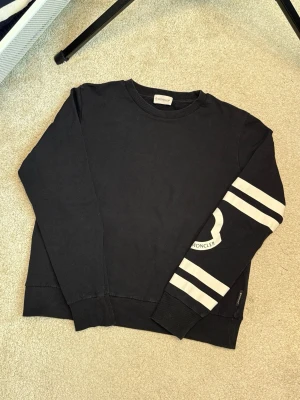 Svart sweatshirt från Moncler - Svart sweatshirt från Moncler med vita ränder och stor Moncler-logga på ena ärmen. Tröjan har rund halsringning, ribbade muddar och en relaxed passform. Perfekt för dig som gillar stilrena och sportiga plagg.