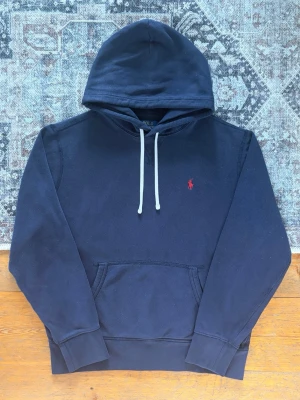 Marinblå Polo Ralph Lauren Hoodie - Storlek M - Hej! 👋 Säljer en klassisk marinblå Polo Ralph Lauren hoodie i storlek M med röd pony-logga. 🐎✨ Tröjan är i mycket gott skick, välvårdad och helt utan hål eller fläckar. Superfräsch! Garanterat 100% äkta med originaltvättlappar kvar (ingen QR-kod på denna årsmodell). Det absolut mest mångsidiga och stilrena basplagget man kan ha i garderoben – passar till allt! Fraktar snabbt! 💯 🚛📦💬 