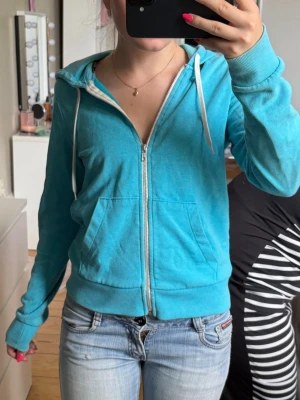 Turkos zip-hoodie med huva - Snygg turkos hoodie med dragkedja framtill och vita snören i huvan. Modellen har två stora fickor fram och ribbade muddar vid ärmar och nederkant🩵