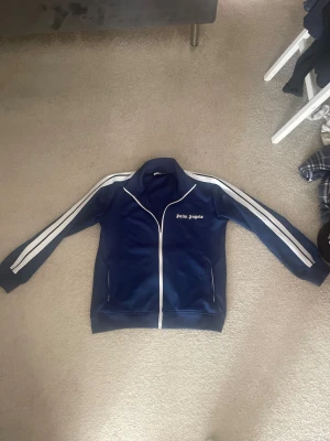 Blå track hoodie från Palm Angels - Säljer nu min Palm angels tröja då jag växt ur den . Det är storlek M och är i fint skick utan skador . Äktenhet vet jag ej men ser bra ut i material osv . För mer frågor är det bara skicka ett meddelande mvh 😄