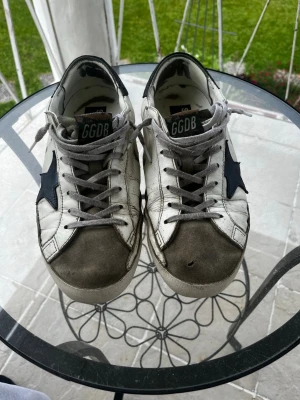 Golden Goose vita sneakers med stjärna - Golden Goose sneakers i vitt läder med grå mockadetaljer på tån och grå skosnören. Svart stjärna på sidan och svart hälparti. Klassisk låg modell med GGDB-logga på plösen och texten SUPER-STAR på insidan. Perfekta för en cool och avslappnad stil. Storlek 43 självklart äkta och kvitto medföljer är hål på båda skorna vid stjärnan men är limmat så syns inte se sista bilden 