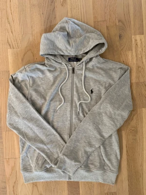Grå zip hoodie från Polo Ralph Lauren - Snygg gråmelerad hoodie från Polo Ralph Lauren med dragkedja framtill och klassisk huva med vita snören. Ikoniska mörkblå loggan broderad på bröstet. Tillverkad i mjukt bomullsmaterial och har ribbade muddar vid ärmslut och nederkant.