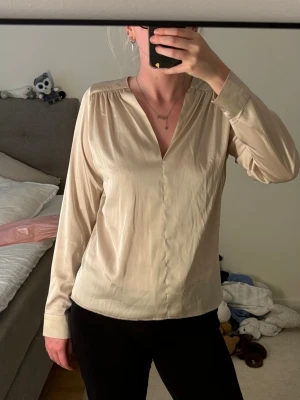Beige v-ringad blus med långa ärmar - Stilren beige blus med v-ringning och långa ärmar. Blusen har diskreta axelveck och manschetter med knappdetalj. Tillverkad i ett mjukt, lätt material som ger ett snyggt fall. Perfekt att matcha med både jeans och kjol.