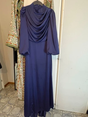 Lång marinblå klänning med drapering - Elegant långklänning i marinblått med vackert draperad framsida och långa puffärmar. Klänningen har en klassisk, flytande siluett och är tillverkad i ett glansigt material som ger en lyxig känsla. Perfekt för speciella tillfällen. Har ett litet hål nere går att fixa lätt.