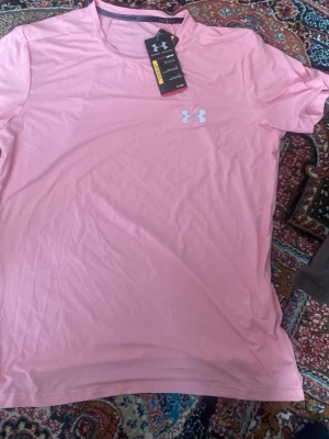  Under Armour t shirt  - Snygg rosa tränings-tshirt från Under Armour med klassisk logga i vitt på bröstet. Tillverkad i ett lätt och snabbtorkande funktionsmaterial som passar perfekt till gymmet eller löprundan. Rund halsringning och korta ärmar för maximal rörelsefrihet.