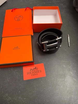 Svart Her skärp med silver H-spänne - Snyggt svart skärp från Her med ikoniskt H-spänne i silverfärg. Skärpet är gjort i skinn och har vita kontrastsömmar längs kanterna. Perfekt accessoar för att lyfta vilken outfit som helst. Levereras med originalförpackning och Her-kort.
