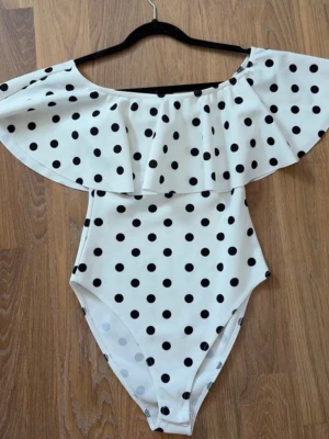 Vit baddräkt med svarta prickar - Supersnygg vit body med svarta prickar och volang över axlarna. Off shoulder-modell som ger en trendig look. Perfekt för dig som gillar retrostil och vill sticka ut lite extra. Materialet känns mjukt och stretchigt.