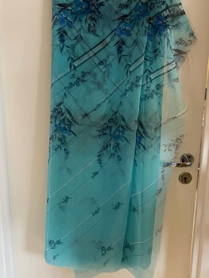 Turkos tyg med blått blommönster - Snygg turkos tyg med stora blå blommor och tunna diagonala ränder. Lätt och luftig i ett tunt, transparent material som ger en fräsch känsla. Perfekt att slänga över axlarna för en extra touch till din outfit. Made in Japan, 113 x 576 cm.