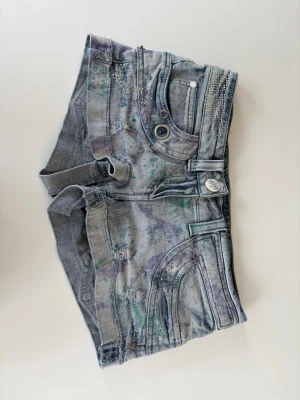 Shorts från River Island  - Unika grå denimshorts från River Island med färgstänk i lila och grönt, dekorerade med små nitar och stenar på fickorna. Shortsen har flera fickor, bälteshällor och råa benslut för en edgy look. Perfekta för dig som gillar detaljer och streetstyle.