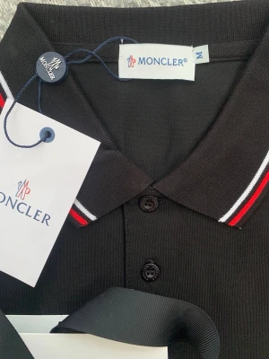 Svart Moncler pikétröja med ränder - Snygg svart pikétröja från Moncler med klassisk krage och knappar framtill. Kragen har röda och vita ränder för en sportig touch och bröstet pryds av Monclers ikoniska logotyp. Perfekt för dig som gillar stilrena och exklusiva plagg.