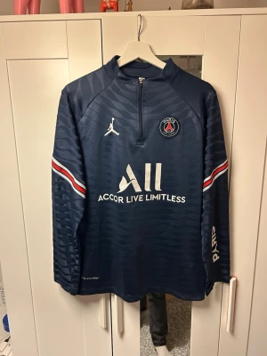 Mörkblå PSG träningströja - Snygg mörkblå långärmad träningströja från PSG och Jordan med halv dragkedja, vita och röda detaljer på ärmarna samt klubbmärke och logga på bröstet. Tillverkad i funktionsmaterial som andas, perfekt för träning eller uppvärmning. Storlek M.