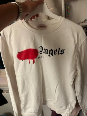 Vit sweatshirt från Palm Angels - Säljer en vit sweatshirt från Palm Angels med svart text och röd spray paint-detalj på bröstet. Tröjan har rund halsringning och är tillverkad i mjukt bomullsmaterial. Perfekt för dig som gillar streetwear och vill sticka ut. Passar storlek M men det står XXL för någon anledning. Den passar mig perfekt och jag bär vanligtvis M