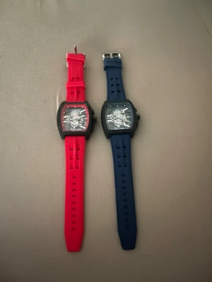 Två sportiga klockor från Richard Mille - Två klockor med transparent urtavla och svart boett. Den ena har rött silikonband och den andra har blått silikonband. Modern design med synlig mekanik och rektangulär form. Perfekt accessoar för dig som gillar en sportig och stilren look.