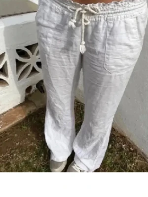 Vita linnebyxor med dragsko - Säljer ett par vita linnebyxor med avslappnad passform och dragsko i midjan. Byxorna har raka ben och stora fickor framtill. Perfekta för en chill och stilren look. Materialet är luftigt linne som känns svalt och skönt.