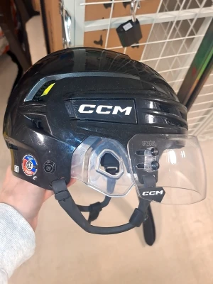 Svart hockeyhjälm CCM med visir - Säljer en svart hockeyhjälm från CCM med genomskinligt visir och justerbart hakband. Hjälmen har tydlig logga på sidan och fram, samt ventilationshål och vadderad insida för extra komfort. Perfekt för dig som spelar ishockey och vill ha bra skydd.