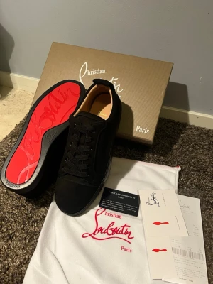 Svarta sneakers från Christian Louboutin - Snygga svarta sneakers från Christian Louboutin med klassisk röd sula och diskret logga. Skorna har rund tå, snörning och är tillverkade i mocka för en lyxig känsla. Perfekta för dig som vill ha en stilren och exklusiv look.