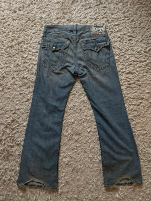 Blå bootcut jeans från True Religion - Säljer ett par blå bootcut jeans från True Religion med klassiska kontrastsömmar och detaljerade bakfickor. Jeansen har låg midja, fem fickor och slitningar vid bensluten. Tillverkade i bomull med en avslappnad passform.