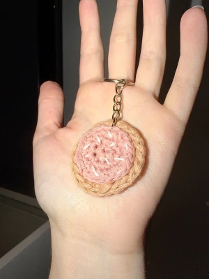 Virkad kaka med glasyr nyckelring - Handgjord virkad nyckelring med rund form i rosa och beige garn. Nyckelringen har en metallkedja och ring för enkel fastsättning på nycklar eller väska. Motiv: kaka med glasyr och strössel!