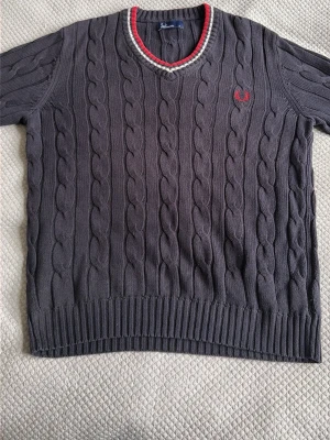 Grå kabelstickad tröja Fred Perry - Kabelstickad grå tröja från Fred Perry med röd och vit detalj vid v-ringningen och röd mudd vid ärmslutet. Tröjan har ett broderat rött Fred Perry-logotyp på bröstet och ribbade muddar. 
