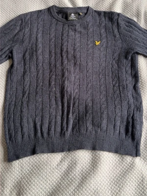 Mörkgrå kabelstickad tröja Lyle & Scott - Mörkgrå kabelstickad tröja från Lyle & Scott med rund halsringning och gul logga på bröstet. Tröjan har ribbade muddar vid ärmslut och nederkant. Perfekt för dig som gillar klassisk och stilren design.