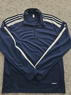 Mörkblå Adidas långärmad träningströja - Mörkblå långärmad träningströja från Adidas med vita ränder längs axlar och ärmar. Tröjan har halv dragkedja framtill, ståkrage och är tillverkad i funktionsmaterial (AEROREADY) som andas. Perfekt för träning eller sportaktiviteter. Aldrig använd.