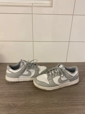Nike Dunk Low grå och vita sneakers - Nike Dunk Low sneakers i vitt och ljusgrått läder med klassisk swoosh på sidan. Lågt skaft, perforerad tåbox och matchande grå snören. Stilren och tidlös modell som passar till det mesta. Perfekt för dig som gillar streetwear och sportig stil.