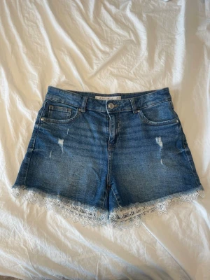 Blå jeansshorts med spetskant - Säljer ett par blå denimshorts  med snygga slitningar och vit spetskant längst ner. Klassisk femficksmodell med knapp och dragkedja framtill. Perfekta för sommaren och ger en cool, feminin vibe.