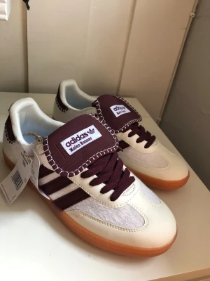 Adidas Wales Bonner sneakers vinröd/vit - Snygga Adidas Wales Bonner sneakers i vitt och vinrött med klassiska tre ränder på sidan, grov sula i gummi och detaljer i mocka och läder. Tungan har Wales Bonner-logga och kontrastsömmar. Perfekta för dig som gillar unik stil och coola detaljer.