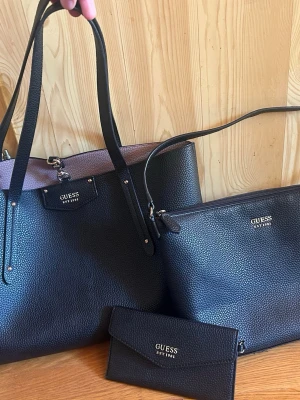 Svart handväska set från Guess - Säljer ett snyggt set från Guess med en stor svart handväska i skinnimitation, matchande axelremsväska och plånbok. Väskorna har guldfärgade detaljer och Guess-logga framtill. Insidan är ljusrosa och väskan har långa handtag. Perfekt för dig som vill ha allt matchat!