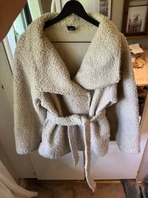 Beige teddyjacka med knytskärp - Mysig beige teddyjacka från Gina Tricot med oversized krage och knytskärp i midjan. Jackan har en fluffig, fårullsliknande yta och är perfekt för kyliga dagar. Stängs med tryckknapp och har en avslappnad passform.