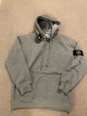Grå hoodie från Stone Island - Snygg grå hoodie från Stone Island med klassisk logopatch på vänster ärm. Hoodien har huva med dragsko, ribbade muddar och magficka. Tillverkad i mjuk bomullsblandning som känns skön mot huden. Perfekt för en avslappnad och trendig look.
