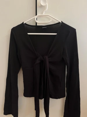 Svart långärmad knytblus - Trendig svart långärmad topp från Gina Tricot med snygg knytning framtill. Modellen är croppad och har en djup v-ringning som ger en cool och modern look. Perfekt att styla med höga jeans eller kjol.