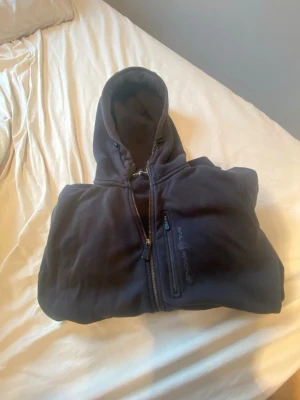 Svart Sail Racing hoodie med dragkedja - Säljer en svart hoodie från Sail Racing med dragkedja framtill och huva. Hoodien har en bröstficka med dragkedja och Sail Racing-loggan broderad på fickan. Tillverkad i mjukt material, perfekt för chill dagar. Klassisk passform och ribbade muddar.