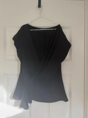 Svart omlottopp med kort ärm - Snygg svart omlottopp med korta ärmar och v-ringad hals. Toppen har en lätt utsvängd passform och är perfekt för att skapa en stilren look. Enkel att matcha med både jeans och kjol.