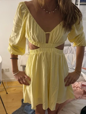 Gul kort klänning med cut-outs - Söt gul kort klänning med v-ringning, cut-outs i midjan och trekvartsärmar. Klänningen har en luftig passform och är tillverkad i ett lätt tyg, perfekt för varma dagar. Resår i midjan ger en snygg siluett och de puffiga ärmarna ger en trendig touch.