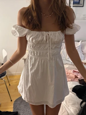 Vit kort sommarklänning med puffärm - Söt vit kort klänning med puffärmar och smockad överdel. Klänningen har snörning framtill och kan bäras off-shoulder eller med ärmarna uppe. Perfekt för varma dagar och har en luftig, lätt känsla.