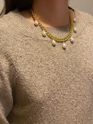 Guldhalsband med pärldroppar - Snyggt halsband i guldfärgad rostfritt med sju droppformade vita pärlor som hänger längs kedjan. Kedjan är lite chunky och ger ett trendigt intryck. Perfekt för dig som vill ha ett statement-smycke med klassiska pärldetaljer.